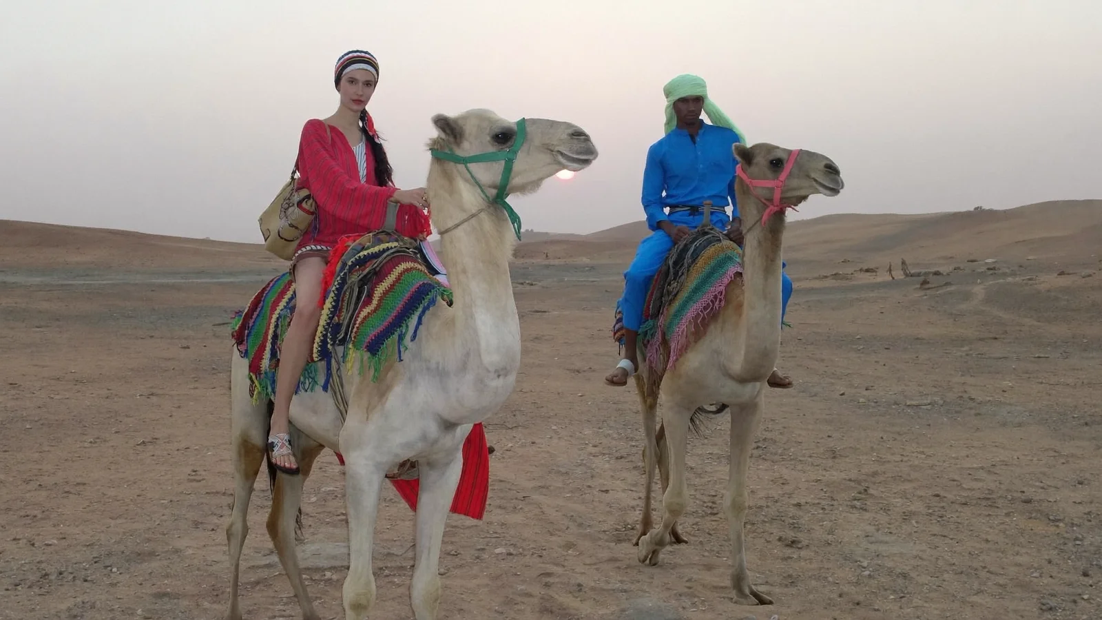 Two riders in red and blue robes on decorated camels in hazy light, quad and camel ride in the Agafay desert