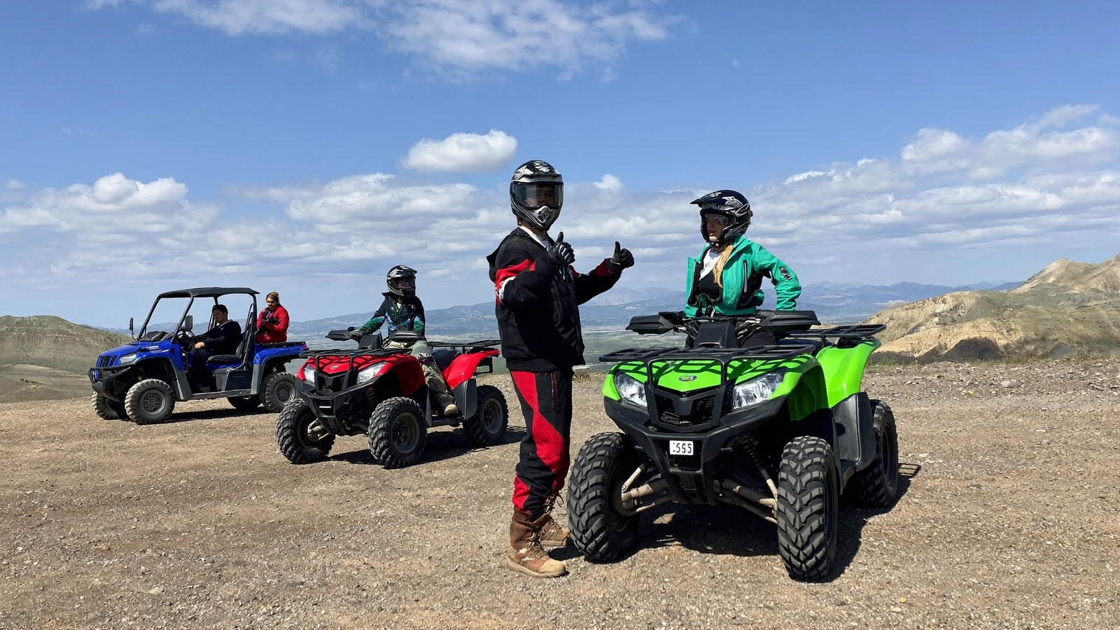Two helmeted riders giving a thumbs up beside a green quad, quad and camel ride in the Agafay desert