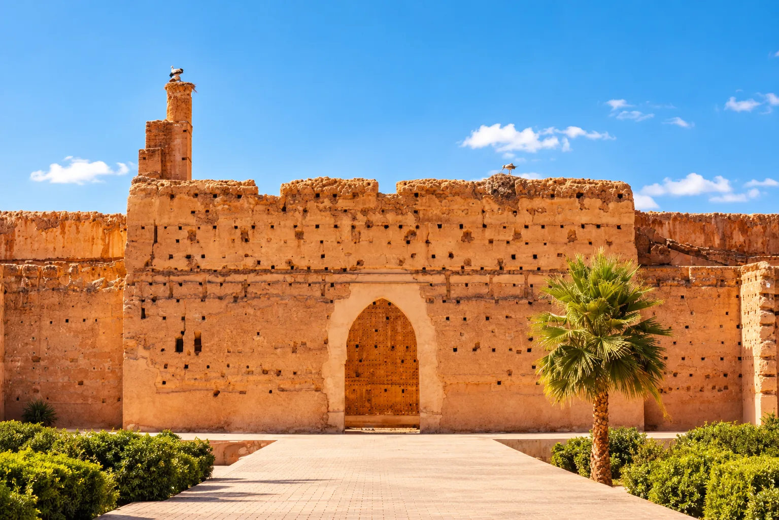 Ochre rampart and arched gate of El Badi Palace with a palm tree and clipped hedges, Marrakech guided tour