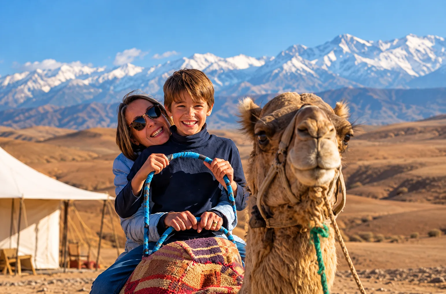 Smiling mother and son on a camel next to a white tent, camel ride in the Agafay desert with snowy Atlas peaks