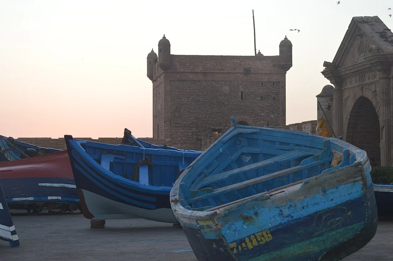 Verwitterte blaue Fischerboote neben einem steinernen Hafenturm unter blassrosa Himmel, Tagesausflug nach Essaouira