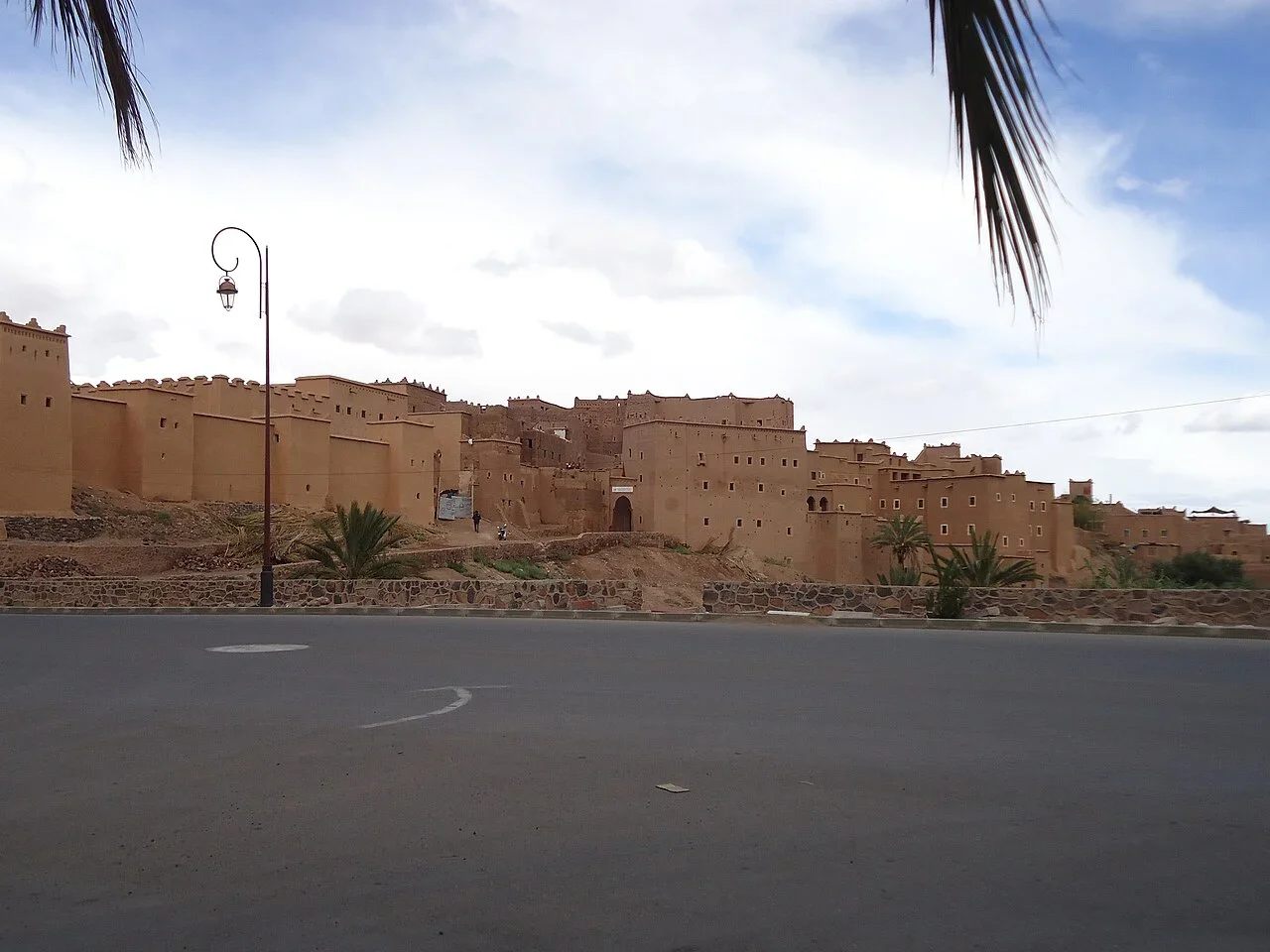 Long earthen kasbah wall beside an empty road with a palm frond in the foreground, Ouarzazate day trip