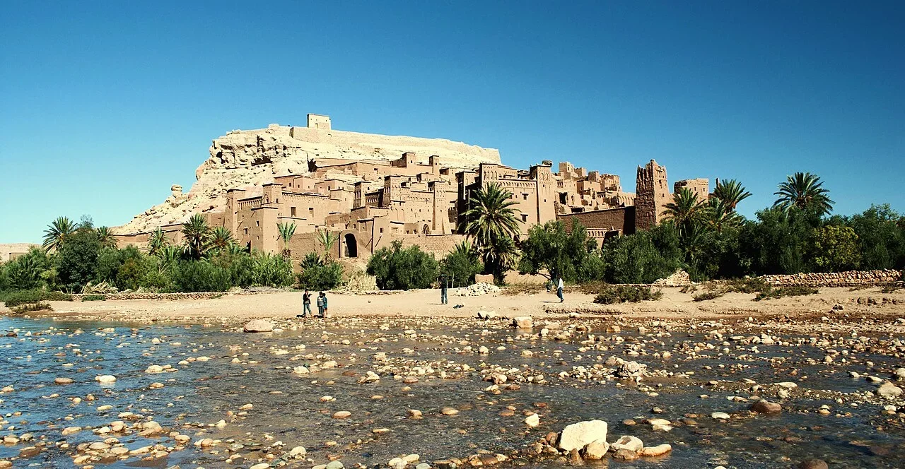 Visitors crossing the shallow riverbed below the ksar and palm trees, Aït Ben Haddou day trip from Marrakech