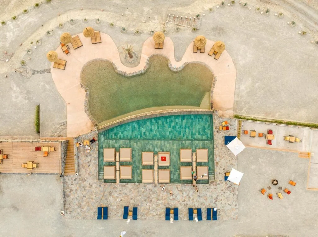 Luftaufnahme eines geschwungenen Pools mit flacher Lagune, Liegen und Schirmen auf Kies, Seven Saints Agafay Pool-Tag