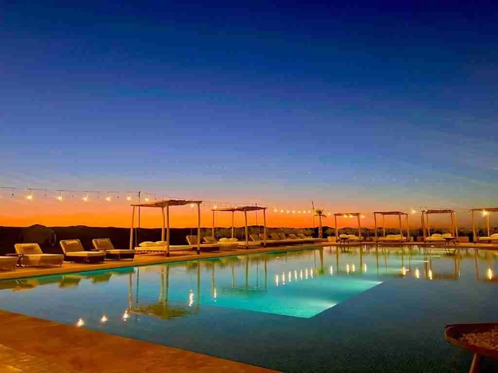 Piscina turchese illuminata che riflette ombrelloni e lucine al calar della notte, day pass in piscina Agafay Valley