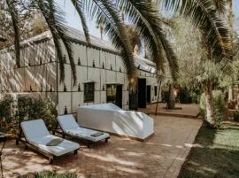 Zwei weiße Sonnenliegen und ein überdachtes Daybed auf einer Ziegelterrasse unter Palmen, Palais Rhoul Pool & Mittagessen