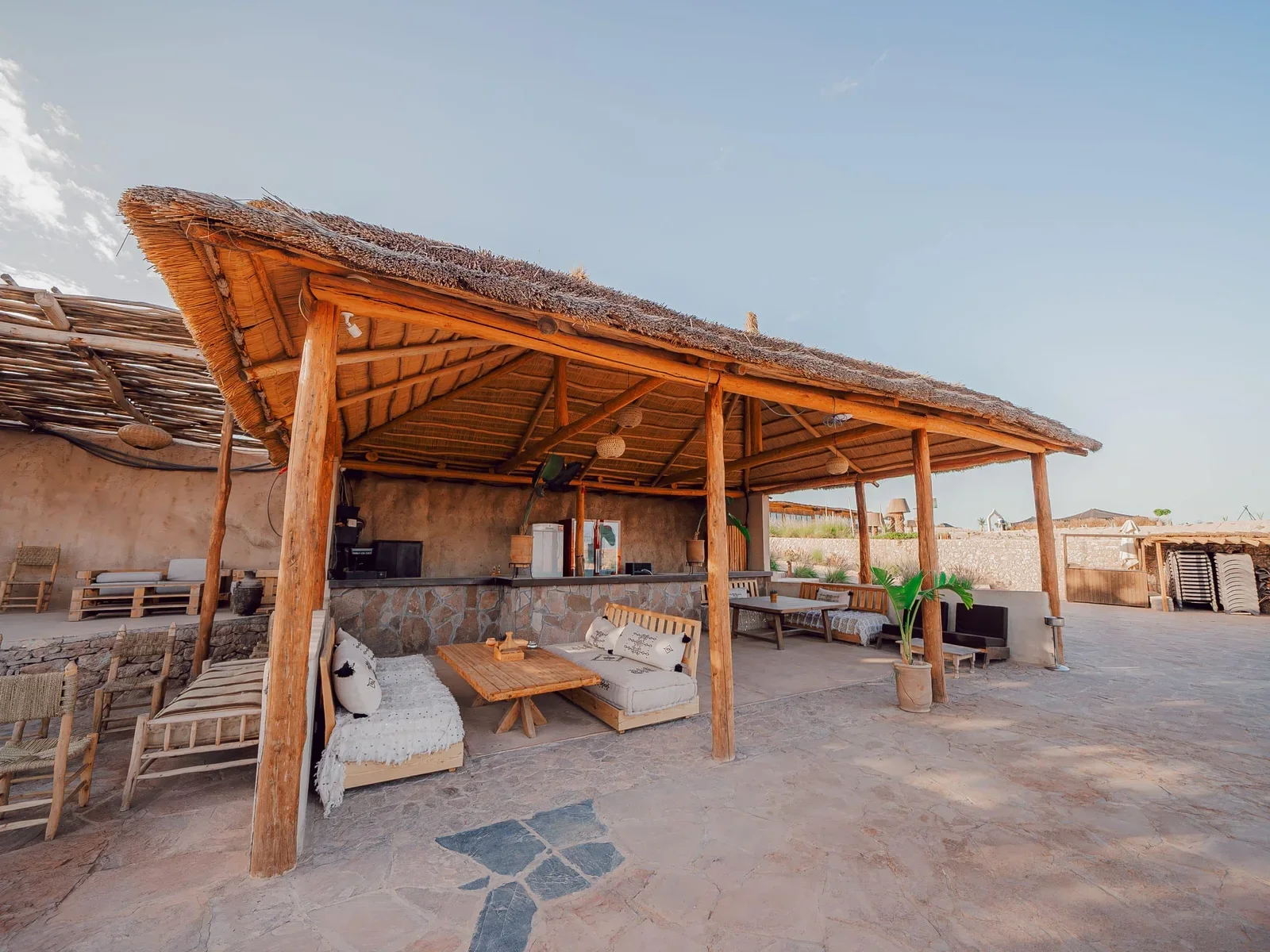 Salón bajo pérgola de paja con sofás y mesa de madera en la terraza de piedra, pase de día con piscina en Le Bedouin Agafay