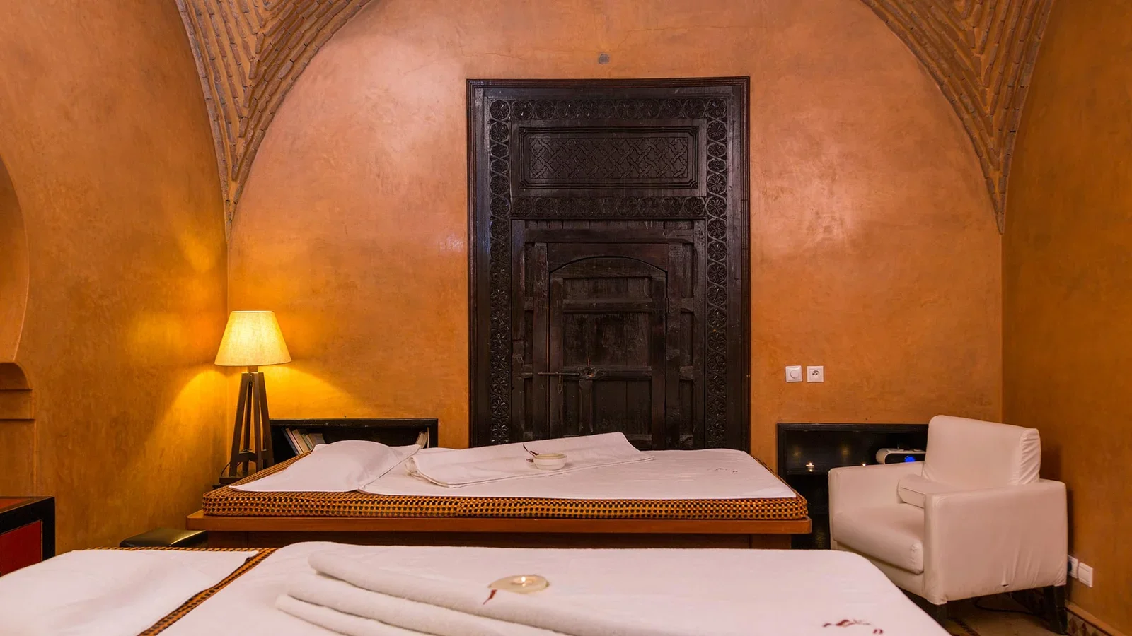 Zwei Massageliegen unter warmem Lampenlicht vor einer geschnitzten Holztür, Spa des Palais Rhoul Pool-Tagespasses